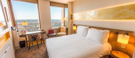 Habitación de hotel con una cama grande, un escritorio, una silla y vistas a la ciudad a través de un amplio ventanal.