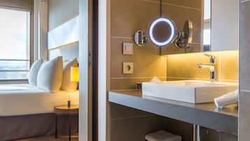 Hotelkamer met een bed, nachtkastje, lamp en een badkamer met wastafel, spiegel en handdoek.