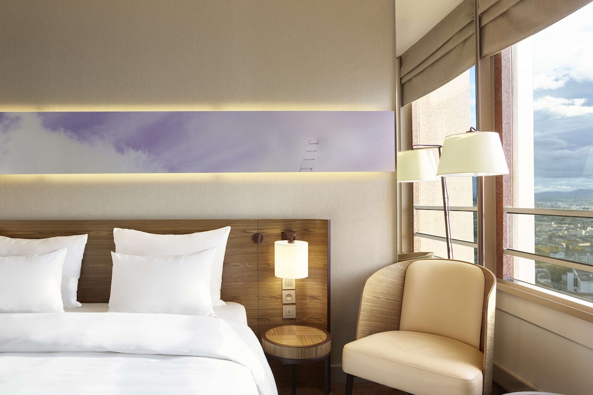 Ein Hotelzimmer mit einem großen Bett, einem runden Beistelltisch, einem Sessel, einer Lampe und Blick auf die Stadt durch das Fenster.