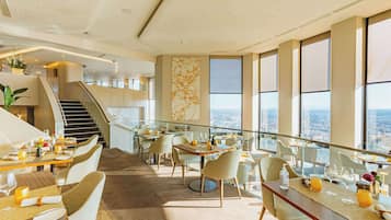 Un espace de restauration moderne avec des tables dressées pour les clients, un escalier et de grandes fenêtres offrant une vue sur la ville.