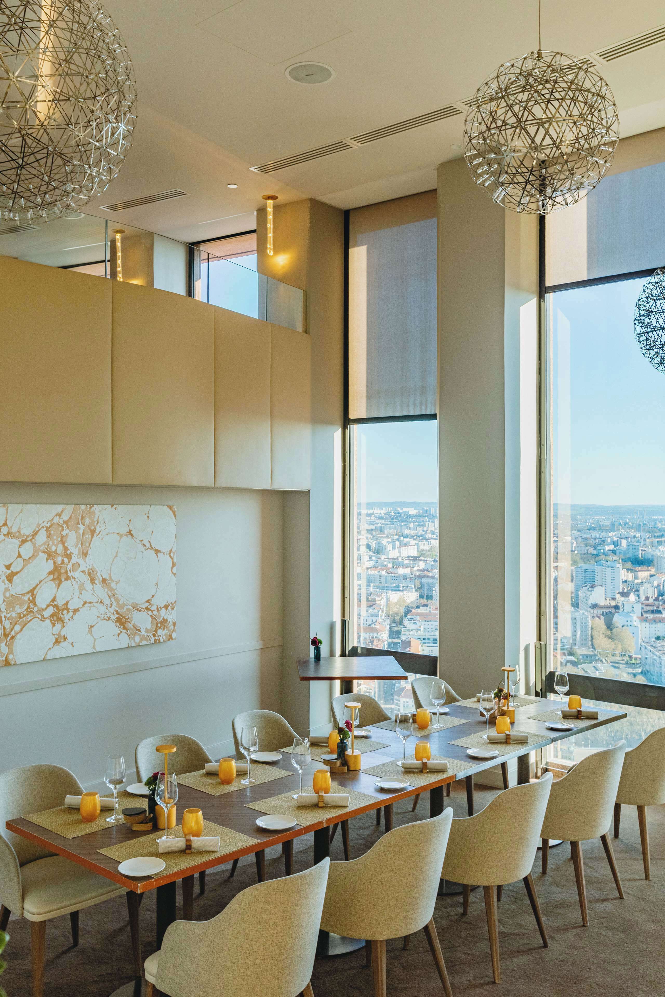 A dining area with a long table set for six, large windows offering a city view, and modern geometric light fixtures.