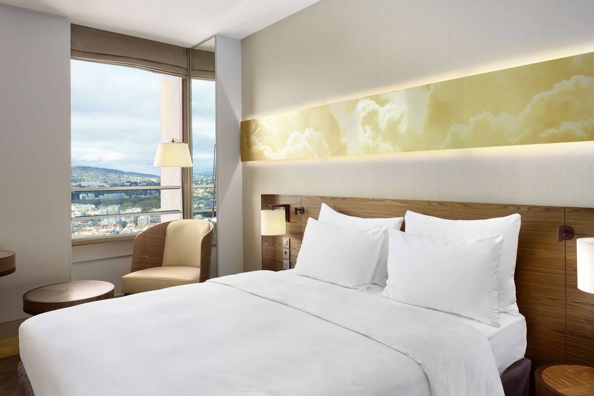 A hotel room with a large bed, a view of the city, a wooden headboard, and a round side table.