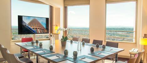 Un salón de conferencias con una gran mesa, sillas, un televisor y ventanas amplias con vistas a la ciudad.