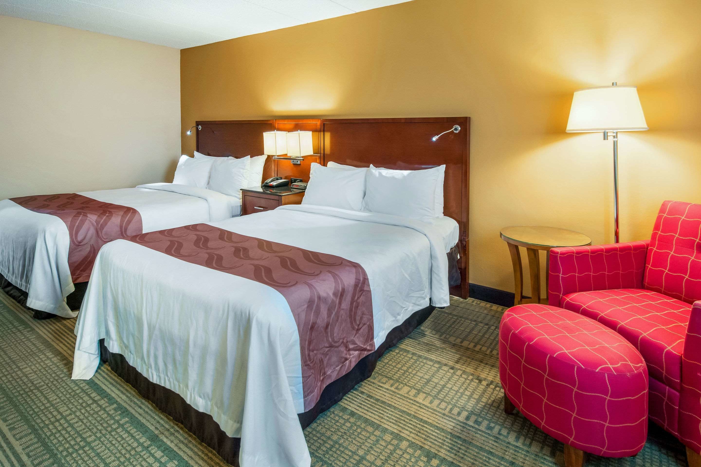 Quarto de hotel com duas camas, uma mesinha, um abajur e uma cadeira vermelha.