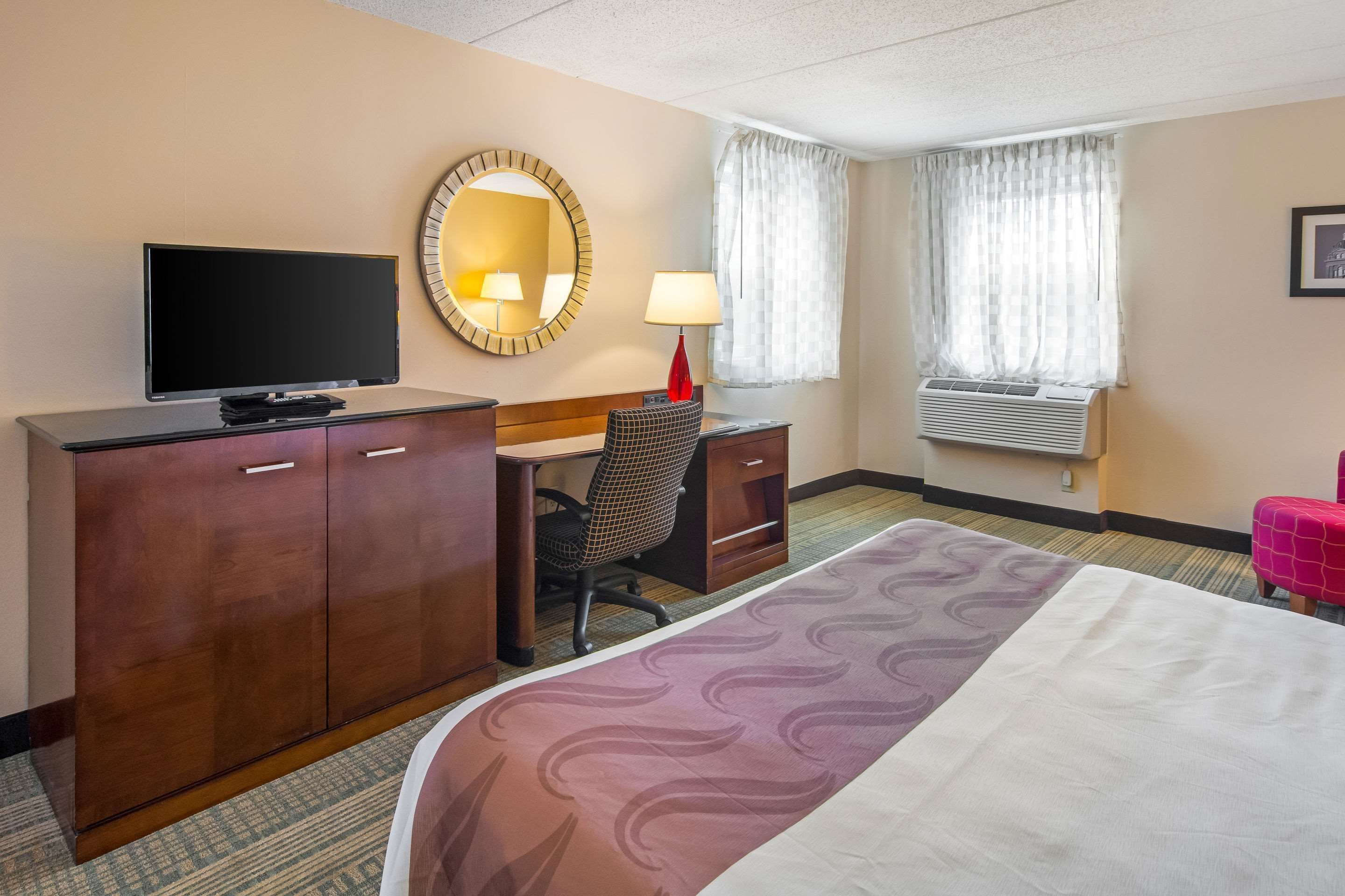 Habitación de hotel con cama, escritorio con silla, televisor, armario, lámpara y ventana con cortinas.