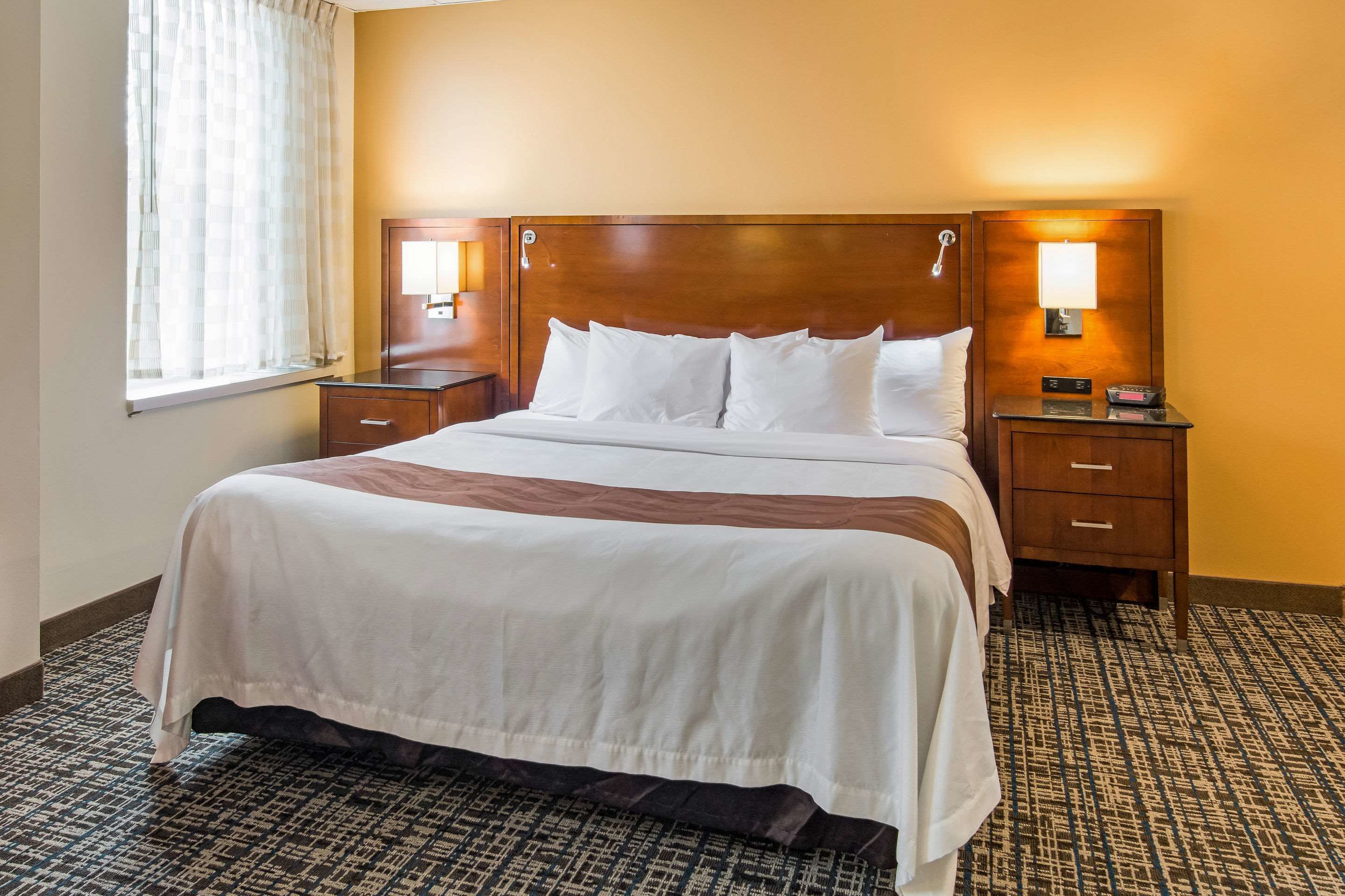 Une chambre d’hôtel avec un grand lit, deux tables de chevet, une petite table et une fenêtre avec des rideaux.