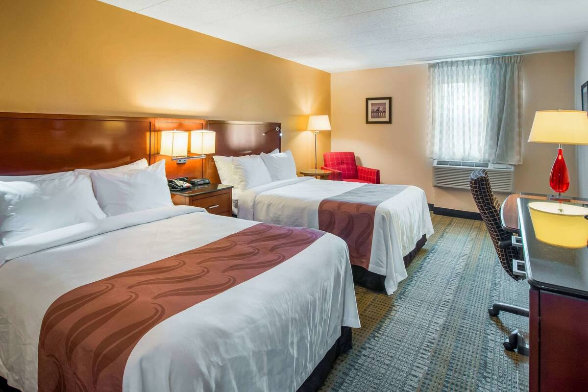 A hotel room with two beds, a desk, a chair, a lamp, and a window with curtains.
