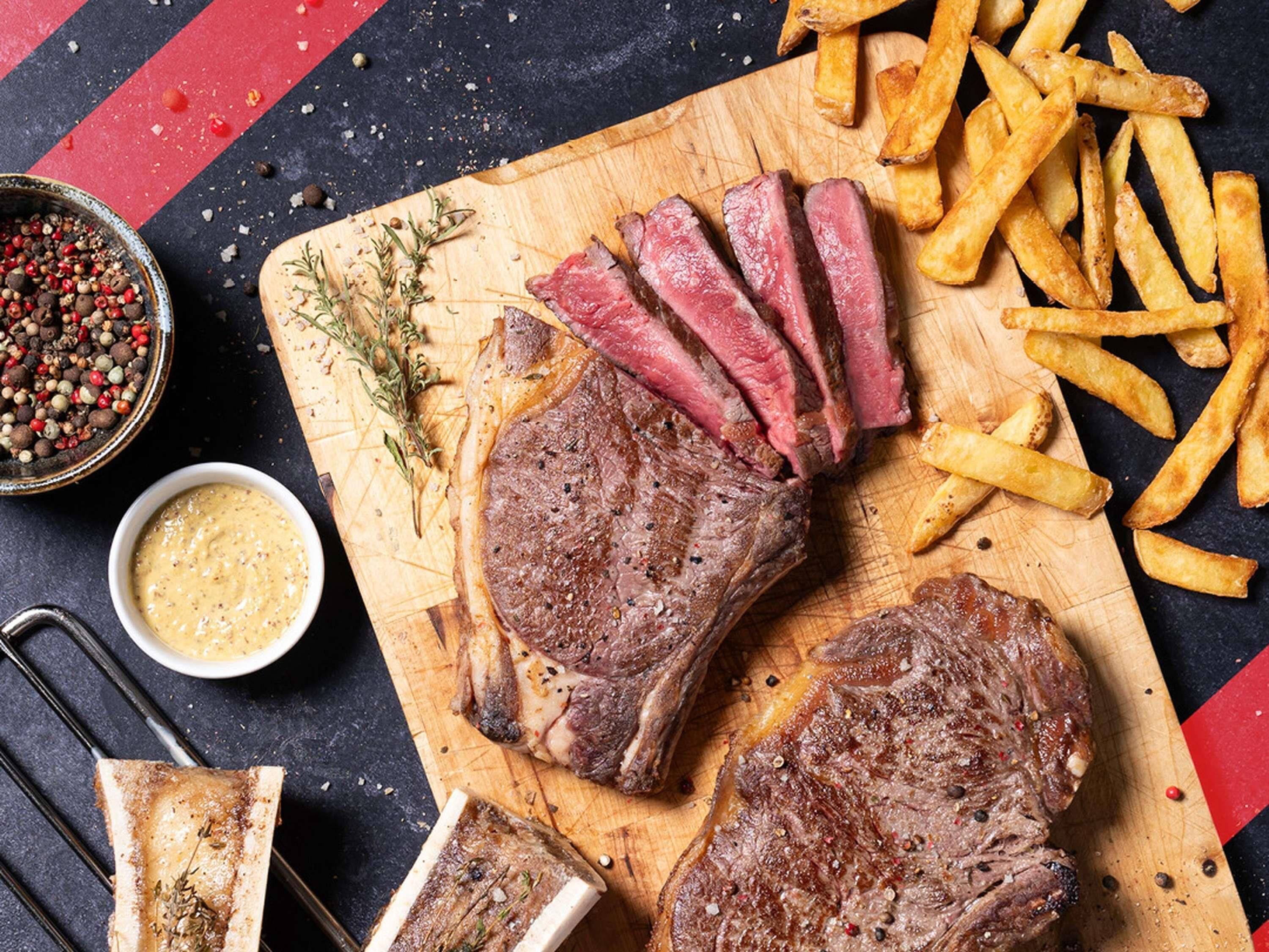 A steak with fries on a wooden board, a bowl of sauce, and a pepper grinder.