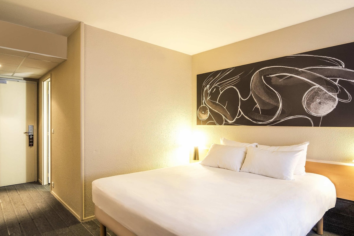 A hotel room with a bed, a wall art piece, a door, and a light fixture.