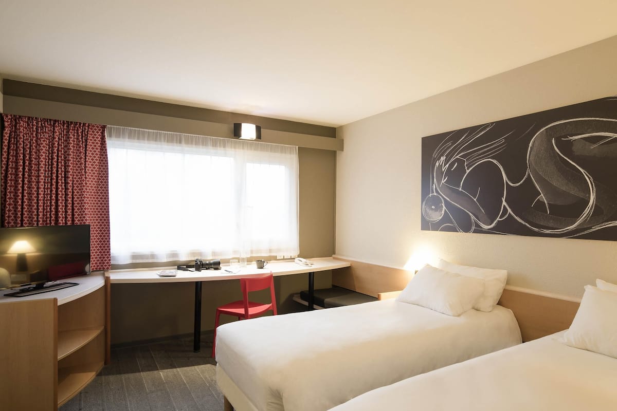 A hotel room with two beds, a desk with a computer, a chair, a window with curtains, and a wall art piece.