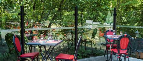 An outdoor patio with tables and chairs, surrounded by lush greenery and a wooden deck.
