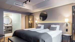 Une chambre d’hôtel moderne avec un grand lit, des tables de chevet, un banc, un miroir rond et une petite salle de bain équipée d’un lavabo et d’un miroir.
