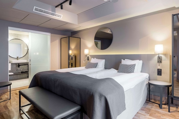 Une chambre d’hôtel moderne avec un grand lit, des tables de chevet, un banc, un miroir rond et une petite salle de bain équipée d’un lavabo et d’un miroir.