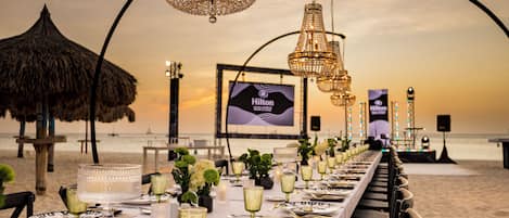 Un montaje para comer frente al mar con una mesa larga, candelabros elegantes y un letrero de Hilton al fondo.
