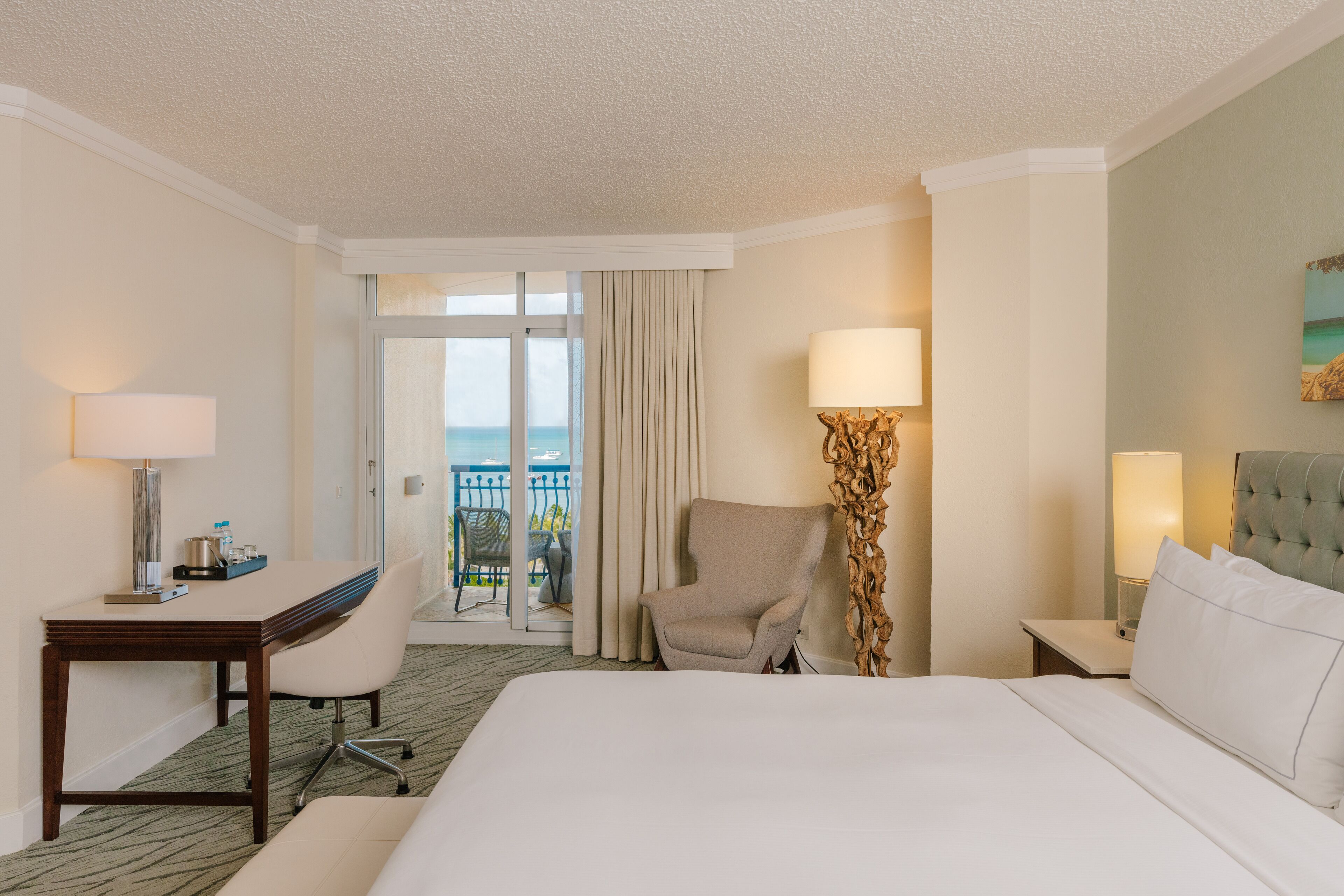 Une chambre d’hôtel comprenant un lit, un bureau avec une chaise, une lampe et une vue sur l’océan, accessible par une porte coulissante.