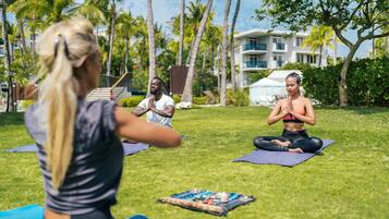 Drei Personen üben Yoga auf Grasmatten im Freien, im Hintergrund Palmen und ein Gebäude.