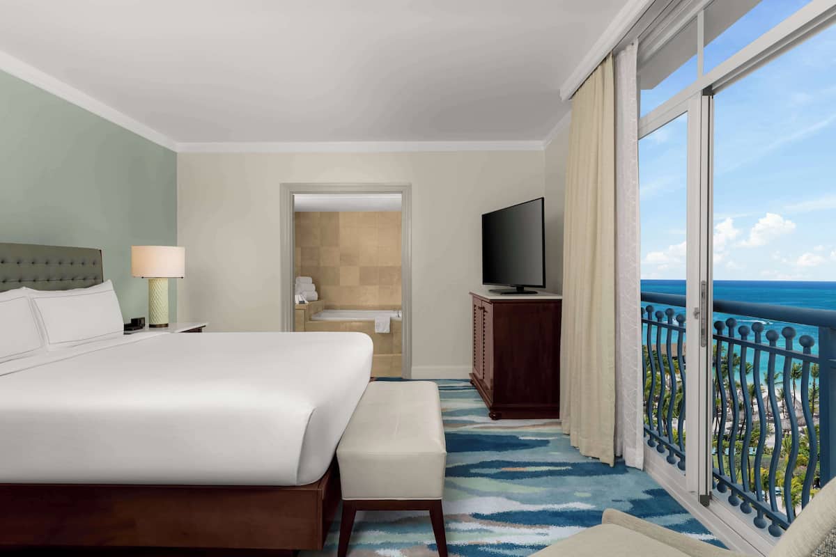 A hotel room with a large bed, a television, a bathroom, and a balcony with a view of the ocean.