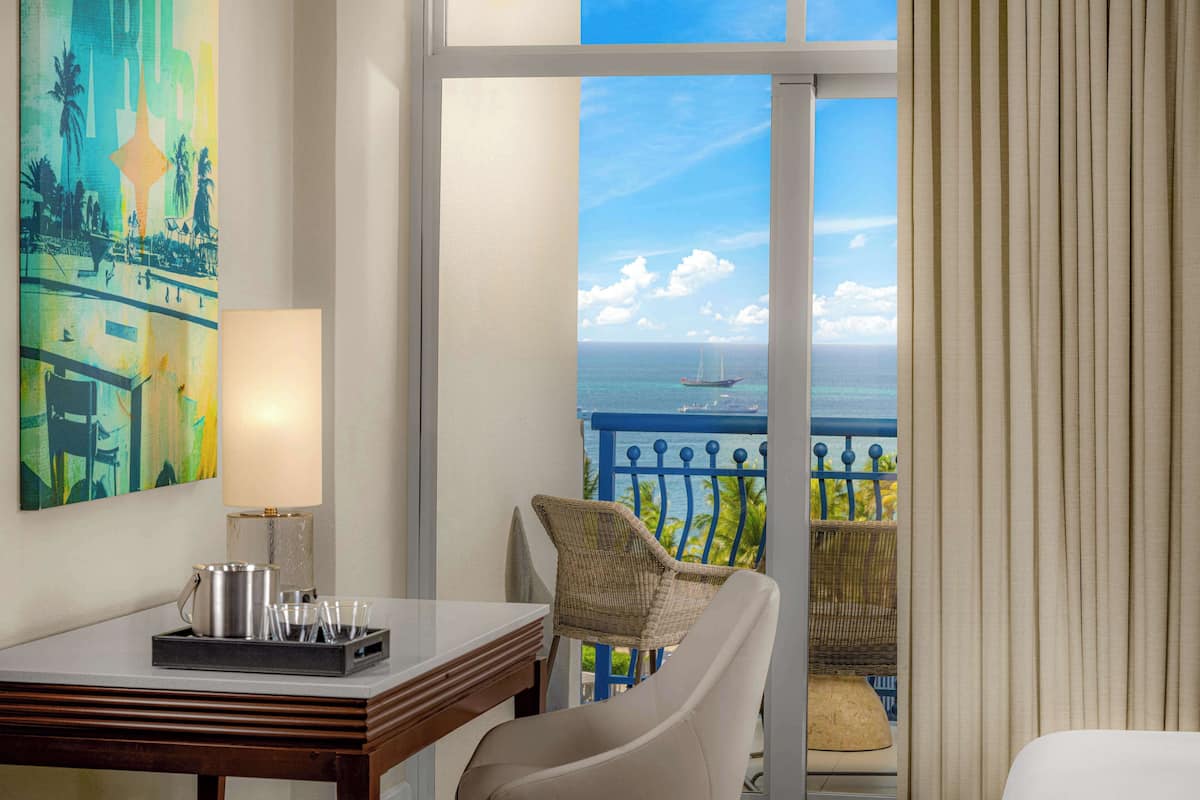 A hotel room with a balcony, a dining table with a lamp, a chair, and a painting on the wall.