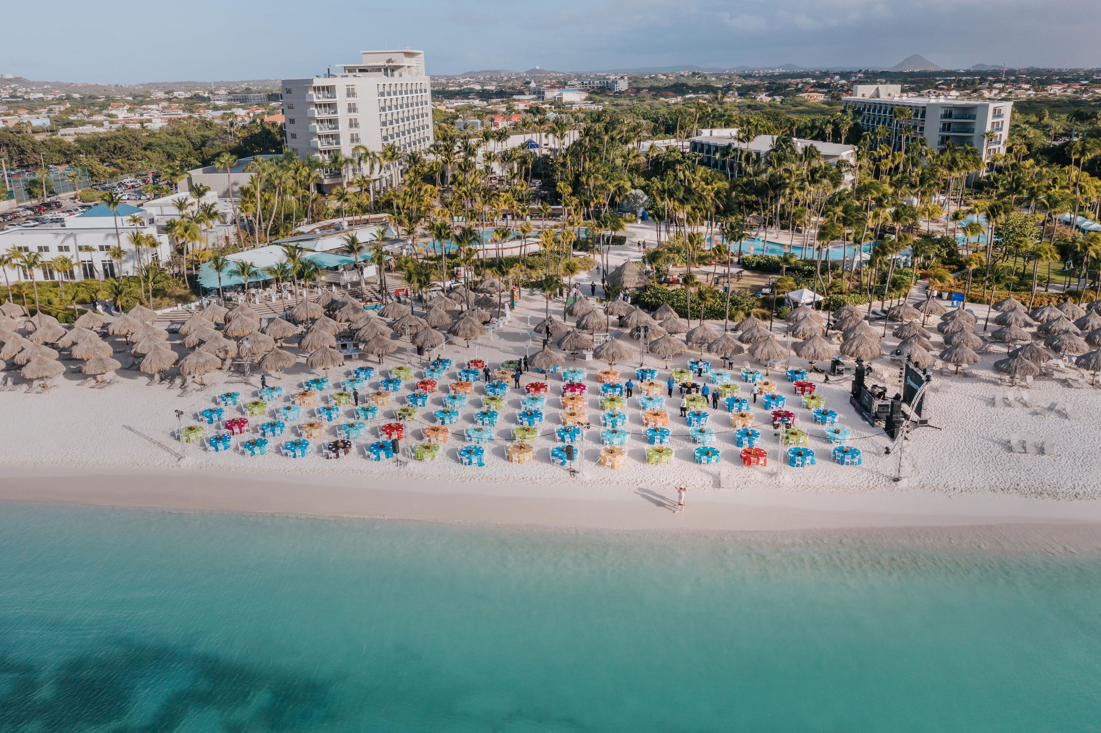 Vista aérea de un complejo turístico en la playa con varias edificaciones, una playa de arena y una vista despejada del océano.
