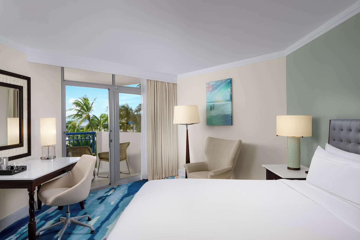 A hotel room with a large bed, a desk, two chairs, a nightstand, and a painting on the wall.