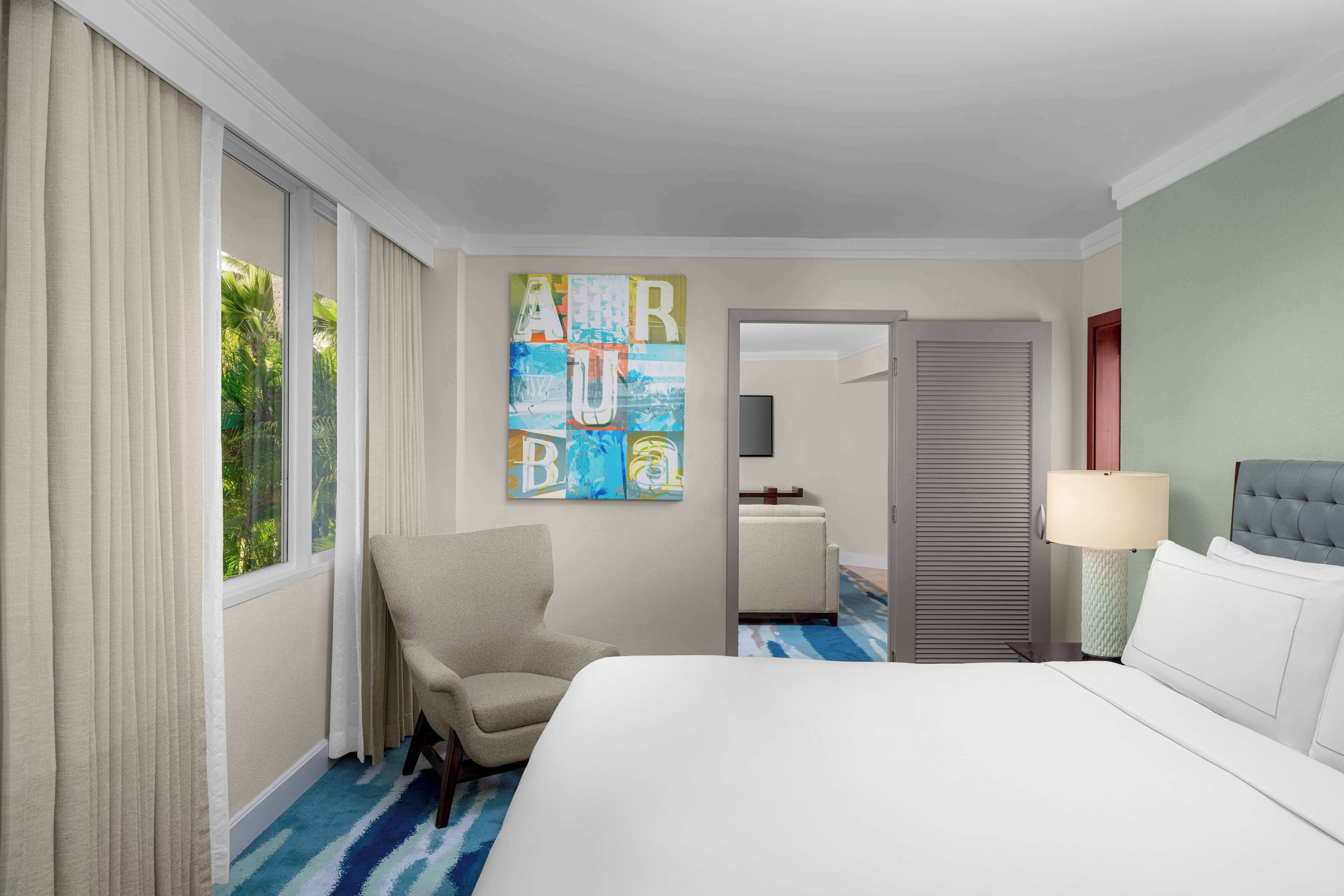 A hotel room with a large bed, a chair, a painting, a lamp, and a view of the outdoors.