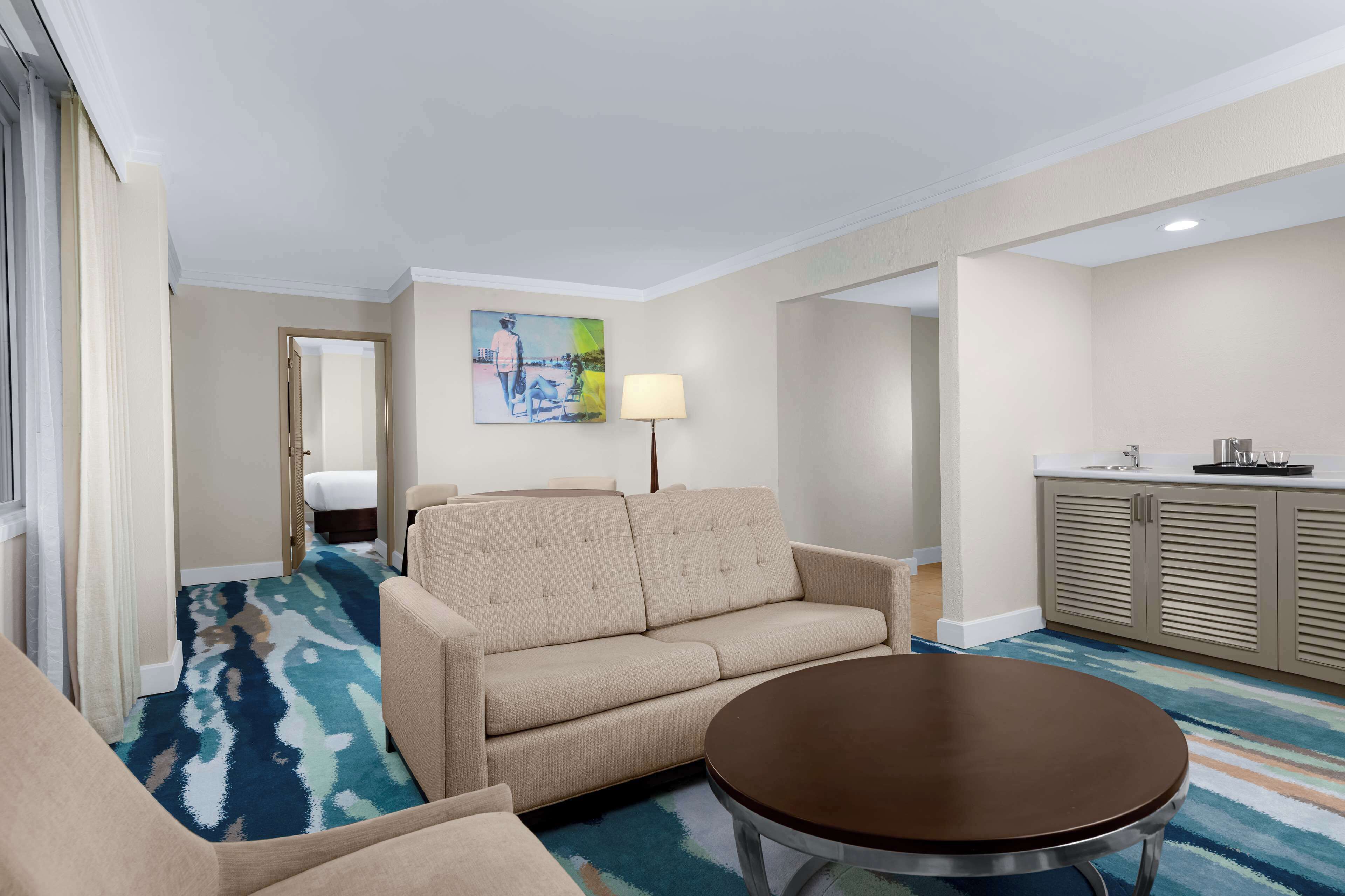 Habitación de hotel con sofá color crema, una mesita redonda, alfombra estampada, un cuadro en la pared y una zona de kitchenette.