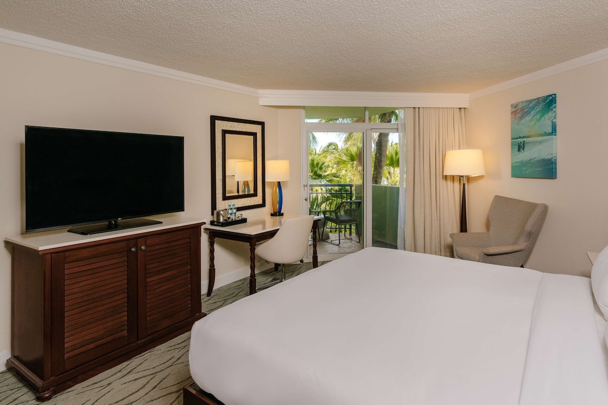 A hotel room with a bed, a desk with a TV, a chair, a lamp, and a balcony with a view of palm trees.