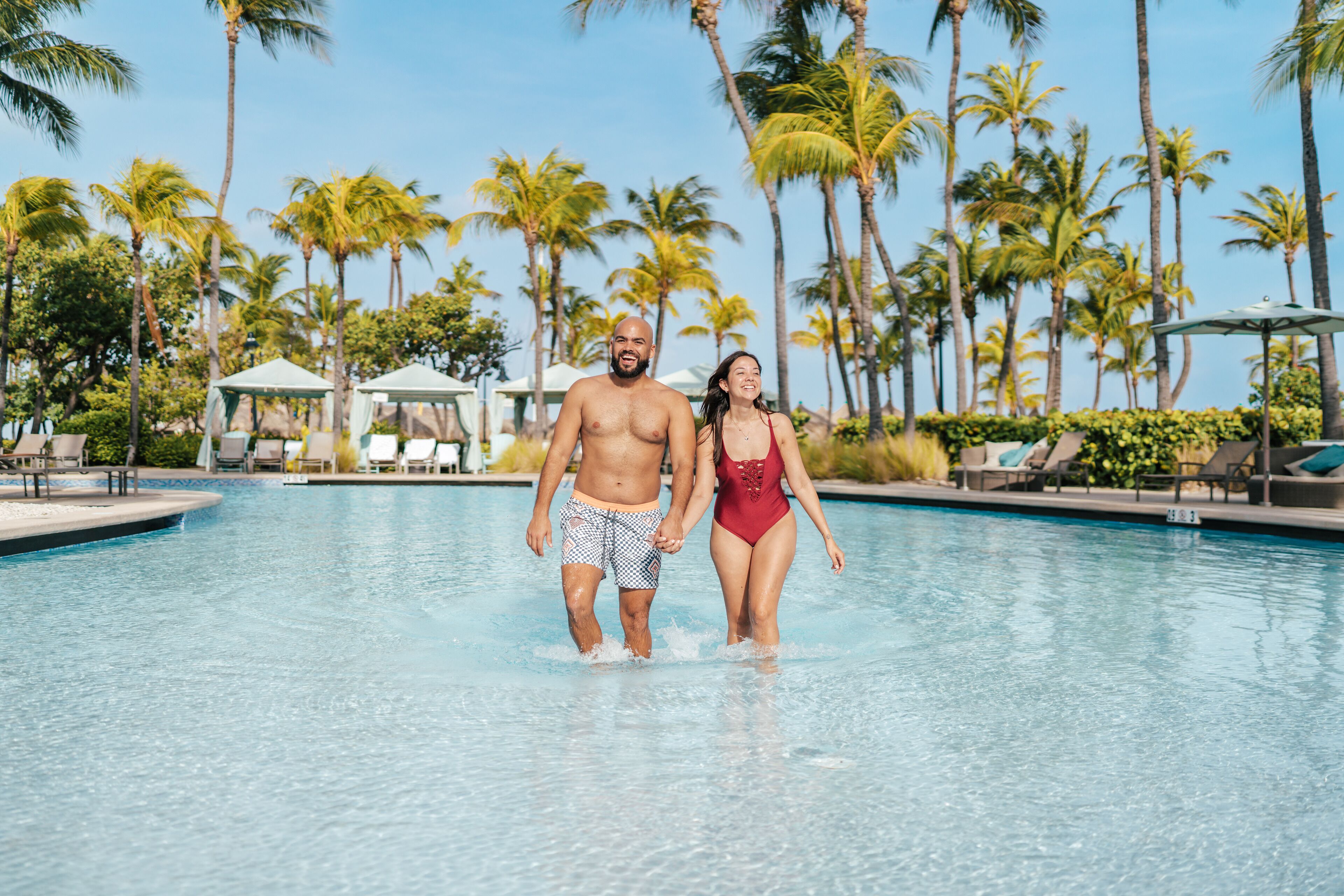 A couple walking in a pool surrounded by palm trees.