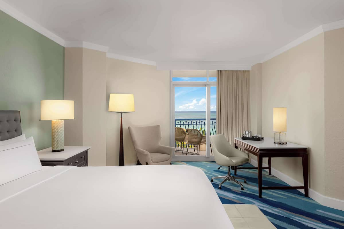A hotel room with a bed, two chairs, a desk, and a view of the ocean through a balcony.