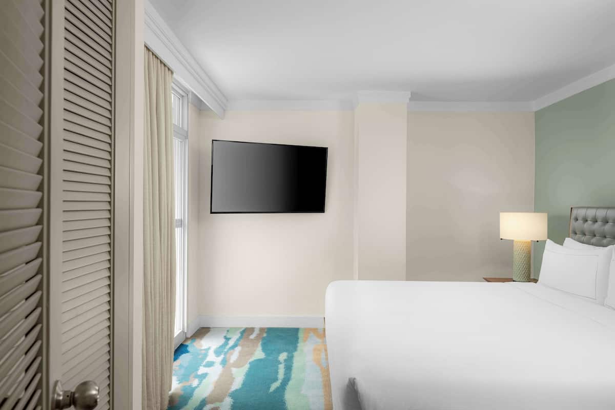 A hotel room with a bed, a television mounted on the wall, a nightstand with a lamp, and a patterned carpet.