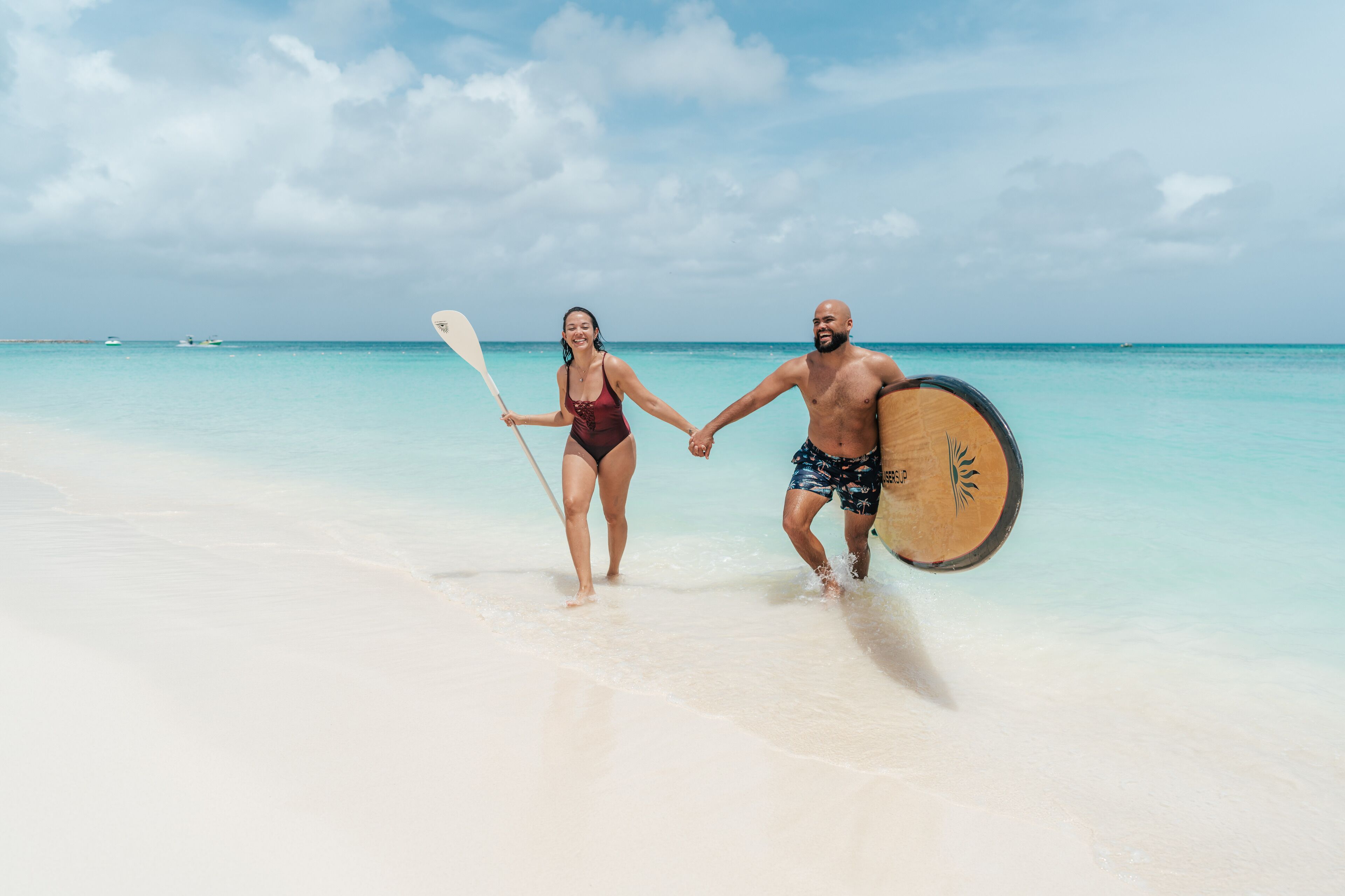 Una pareja camina de la mano por una playa, con el hombre llevando una tabla de remo.