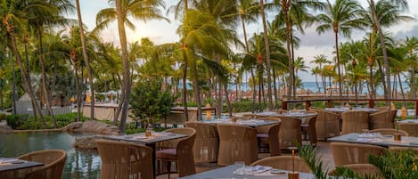 An outdoor dining area with tables and chairs, surrounded by palm trees and a view of the ocean.
