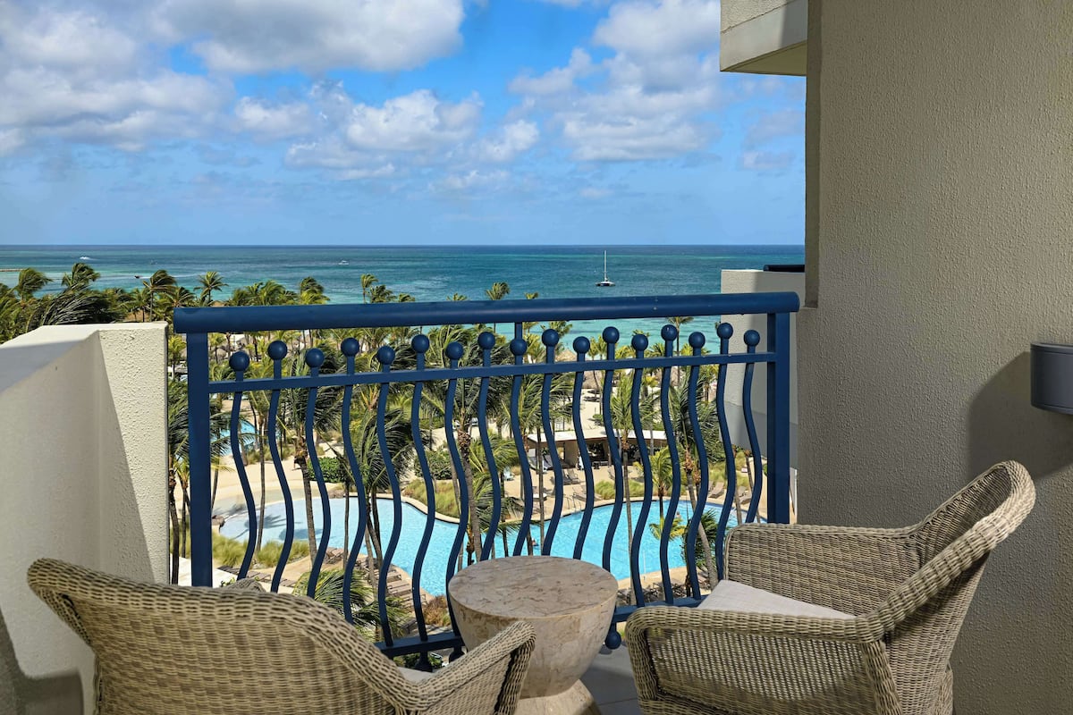 A balcony with wicker chairs and a table, overlooking a pool and beach with palm trees.