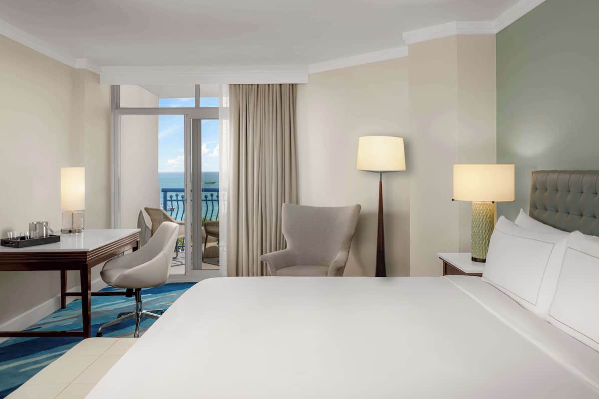 A hotel room with a large bed, a desk, a chair, two bedside lamps, and a view of the ocean through a sliding door.