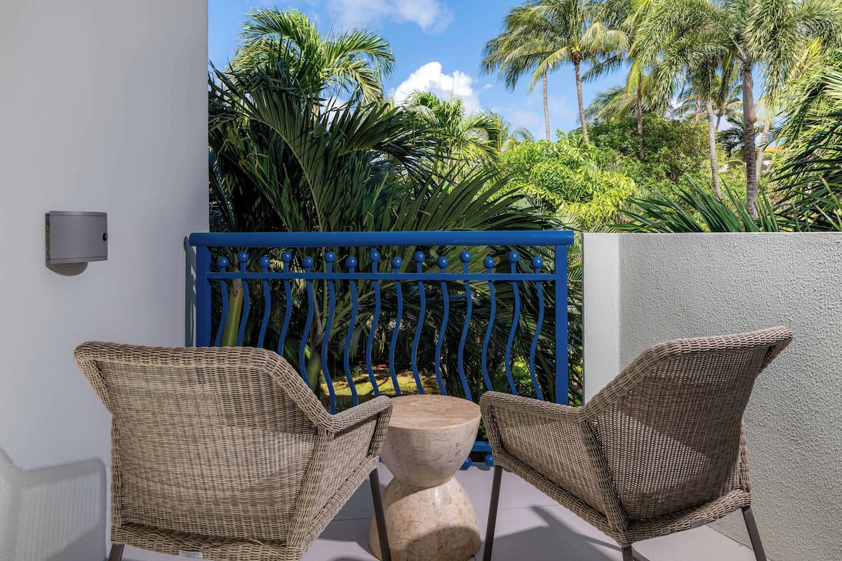 A balcony with wicker chairs and a marble table, surrounded by a blue metal fence and lush greenery.