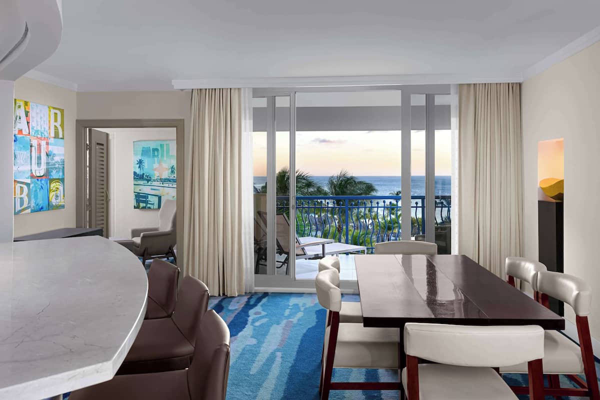 A hotel room with a large dining table, comfortable seating, a view of the ocean, and a balcony with outdoor furniture.