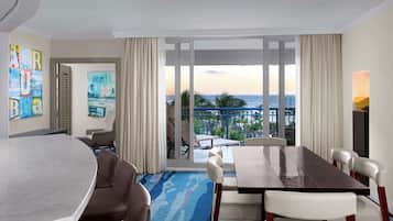 A hotel room with a large dining table, comfortable seating, a view of the ocean, and a balcony with outdoor furniture.