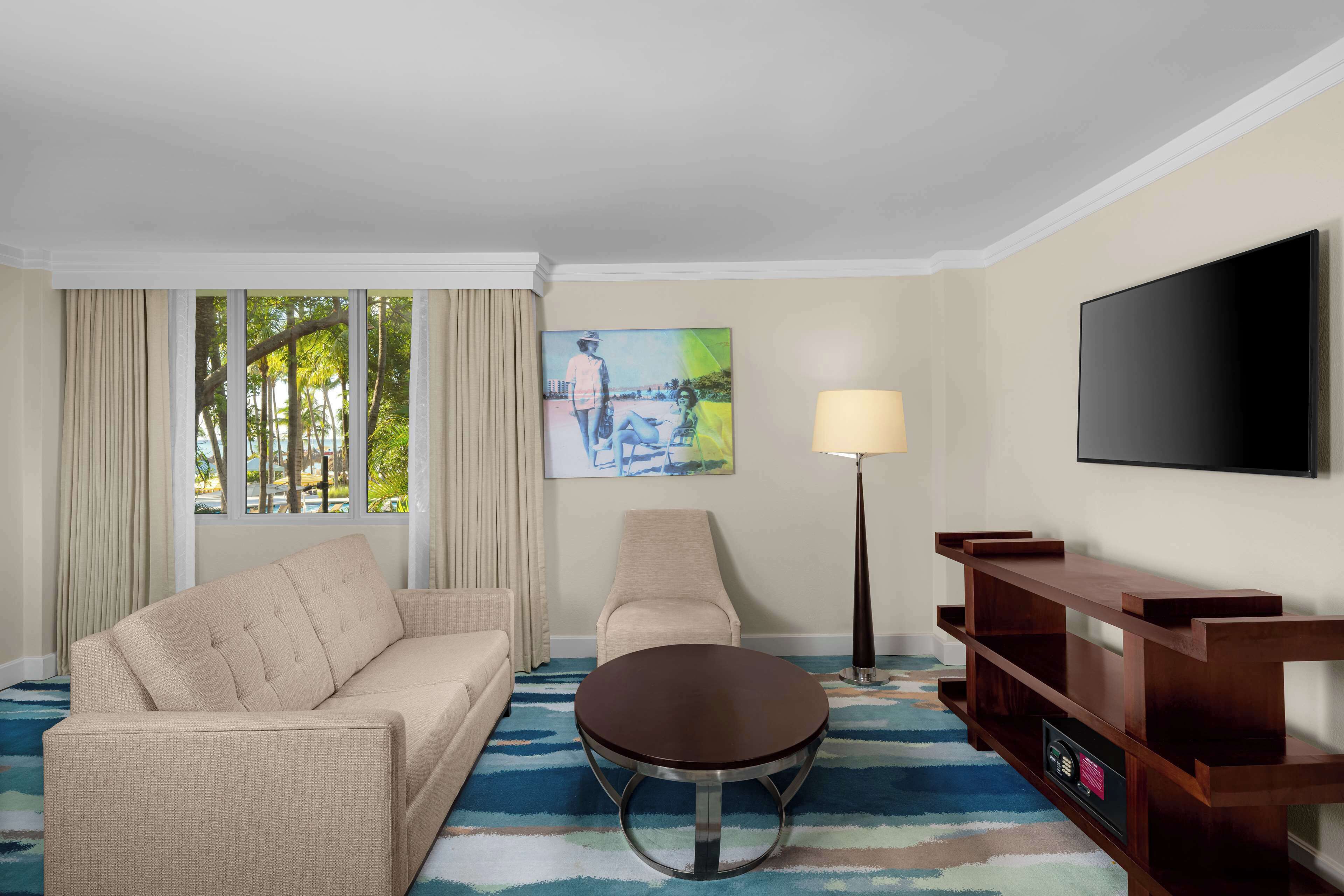 A hotel room with a beige sofa, a round coffee table, a TV on the wall, a painting, and a lamp.