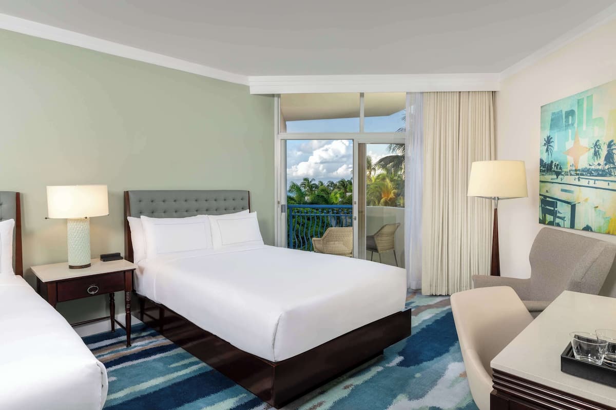 A hotel room with a large bed, two bedside tables with lamps, a chair, a small table with glasses, and a balcony with a view of palm trees.