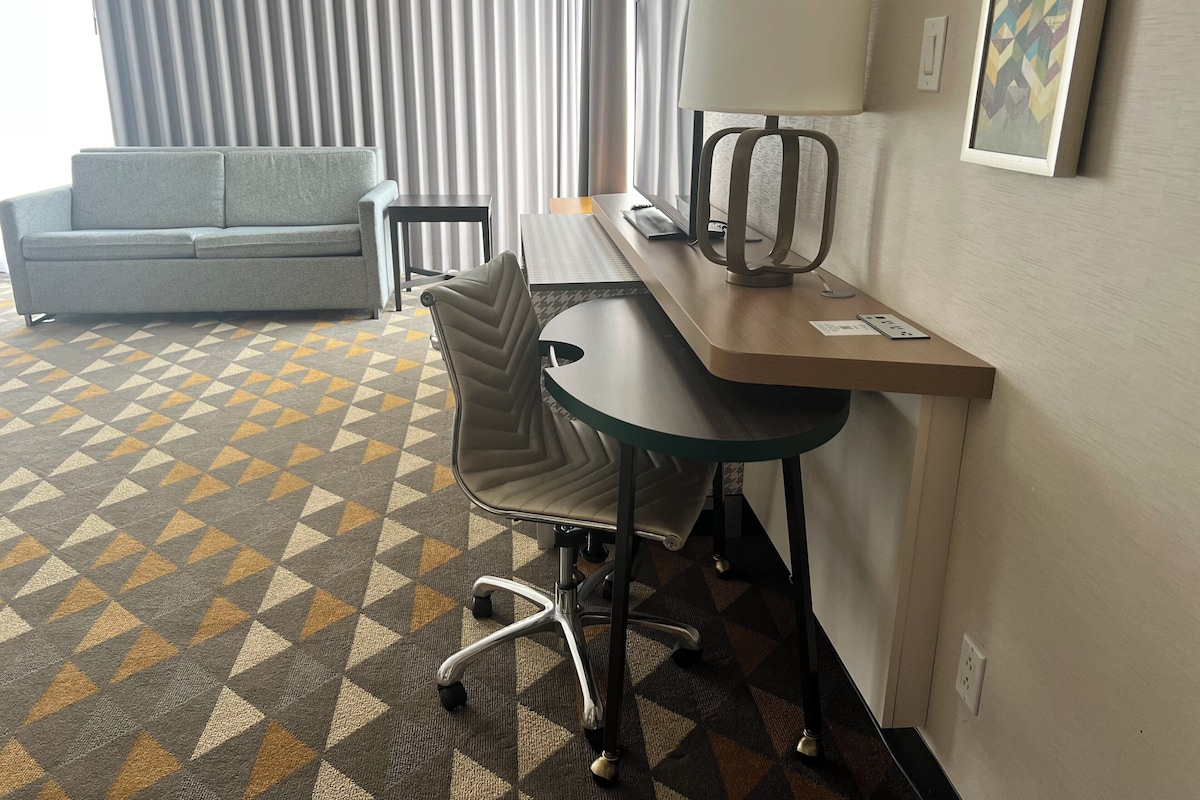 Una habitación de hotel con sofá, escritorio con lámpara, una silla y alfombra estampada.