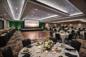 Un salón de baile espacioso con un escenario, varias mesas redondas dispuestas para cenar y una pantalla grande mostrando un mensaje de bienvenida.