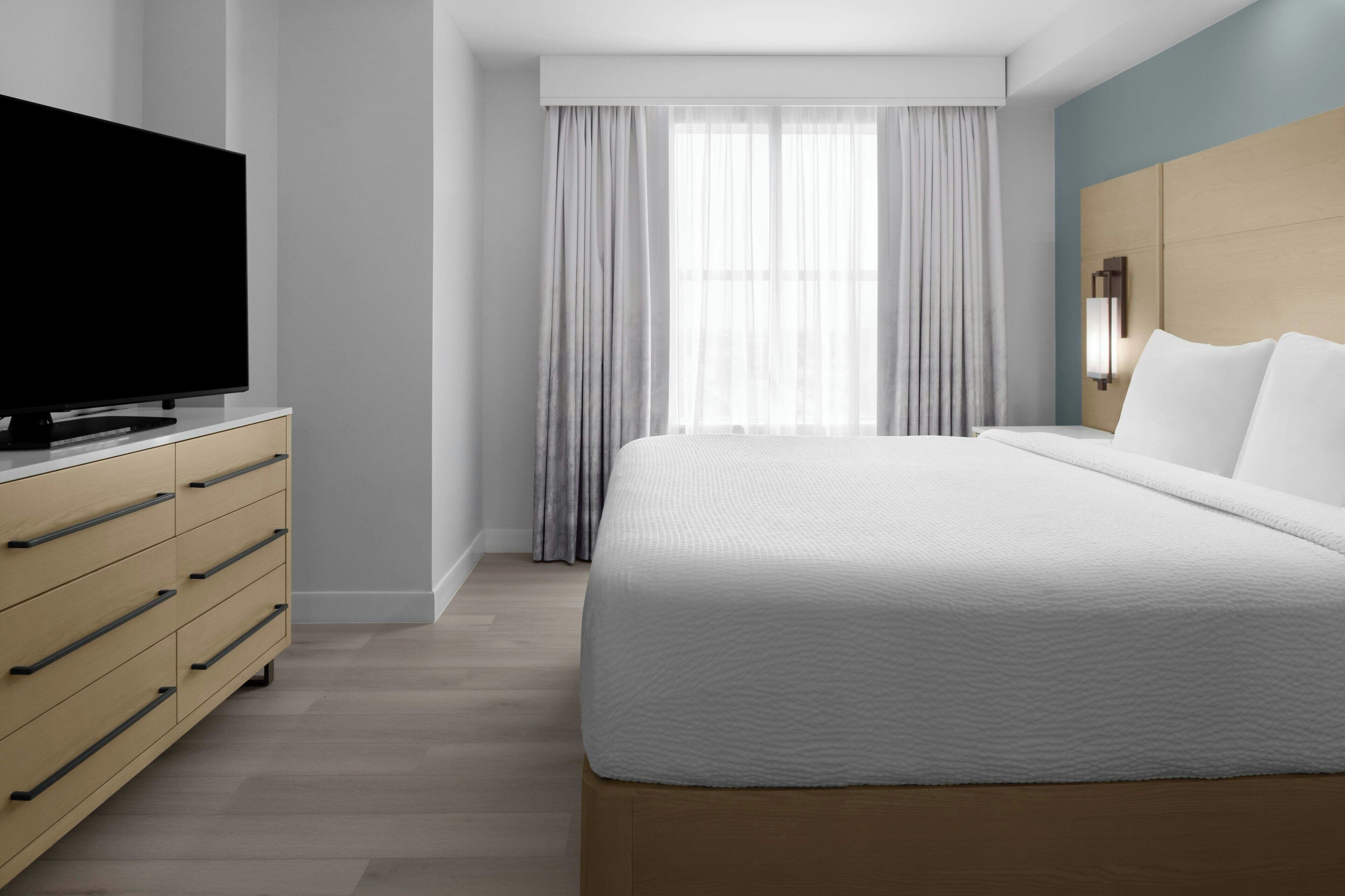 Ein modernes Hotelzimmer mit einem großen Bett, einem Flachbildfernseher auf einer Holzkommode und einem großen Fenster mit transparenten Vorhängen.