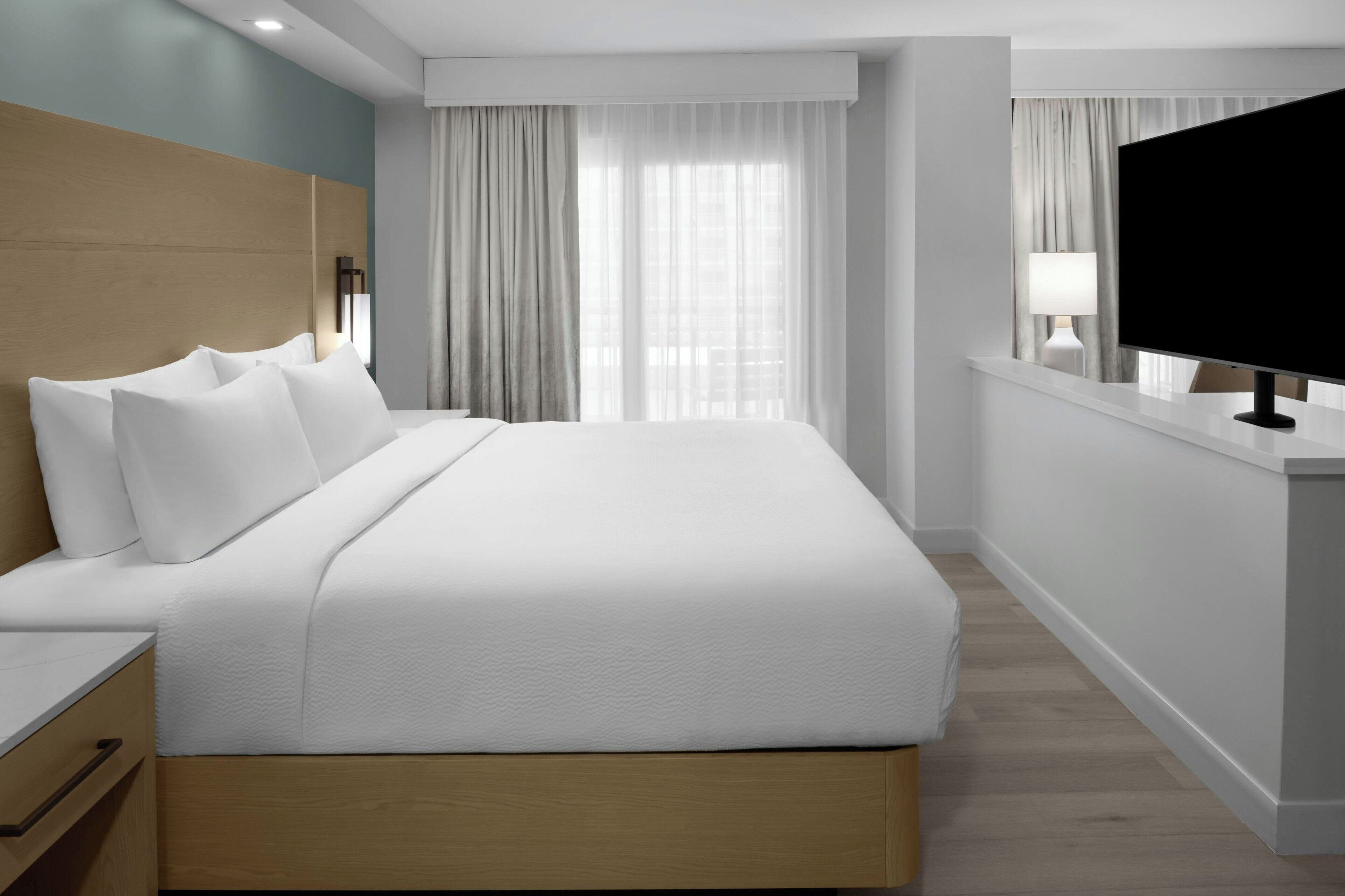 A modern hotel room with a large bed, a flat-screen TV, a bedside table, and a window with curtains.