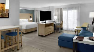 Quarto de hotel com cama, escrivaninha com computador, televisão, sofá e pequena mesa com abajur.