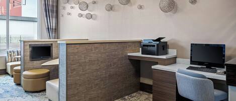 A modern office space with a wooden desk, two monitors, a printer, and a comfortable seating area with ottomans.