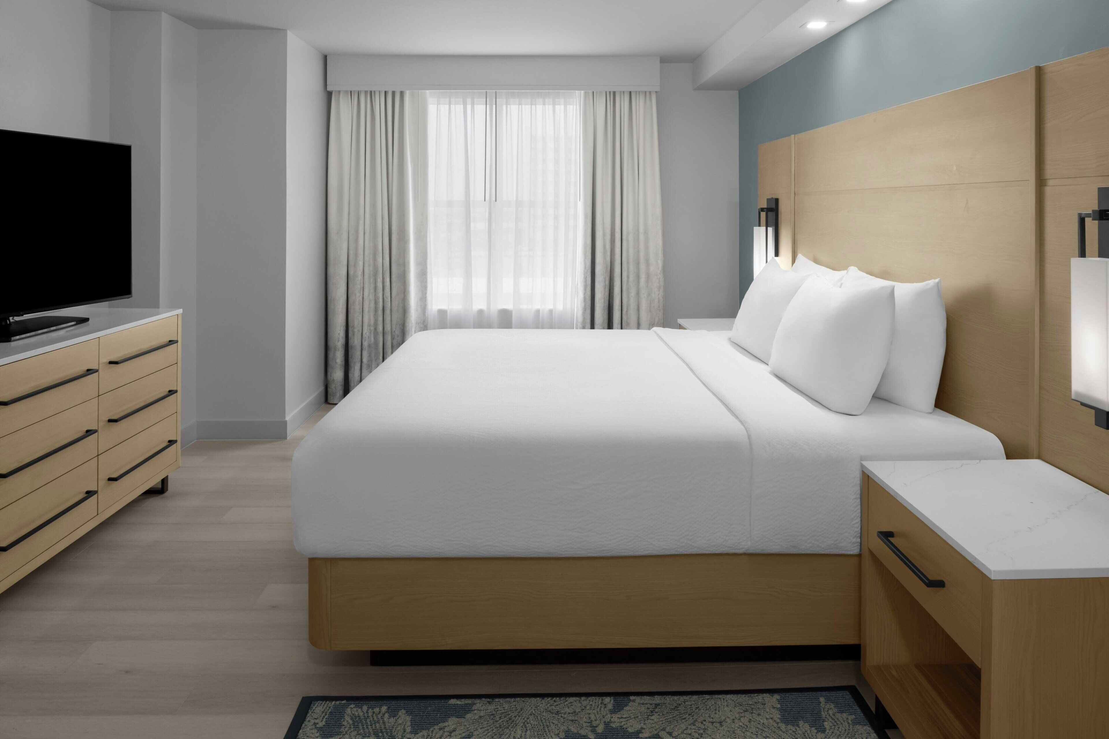 Ein modernes Hotelzimmer mit einem großen Bett, einem Holzkopfstück, einem Flachbildfernseher auf einem Nachttisch und einem großen Fenster mit Vorhängen.