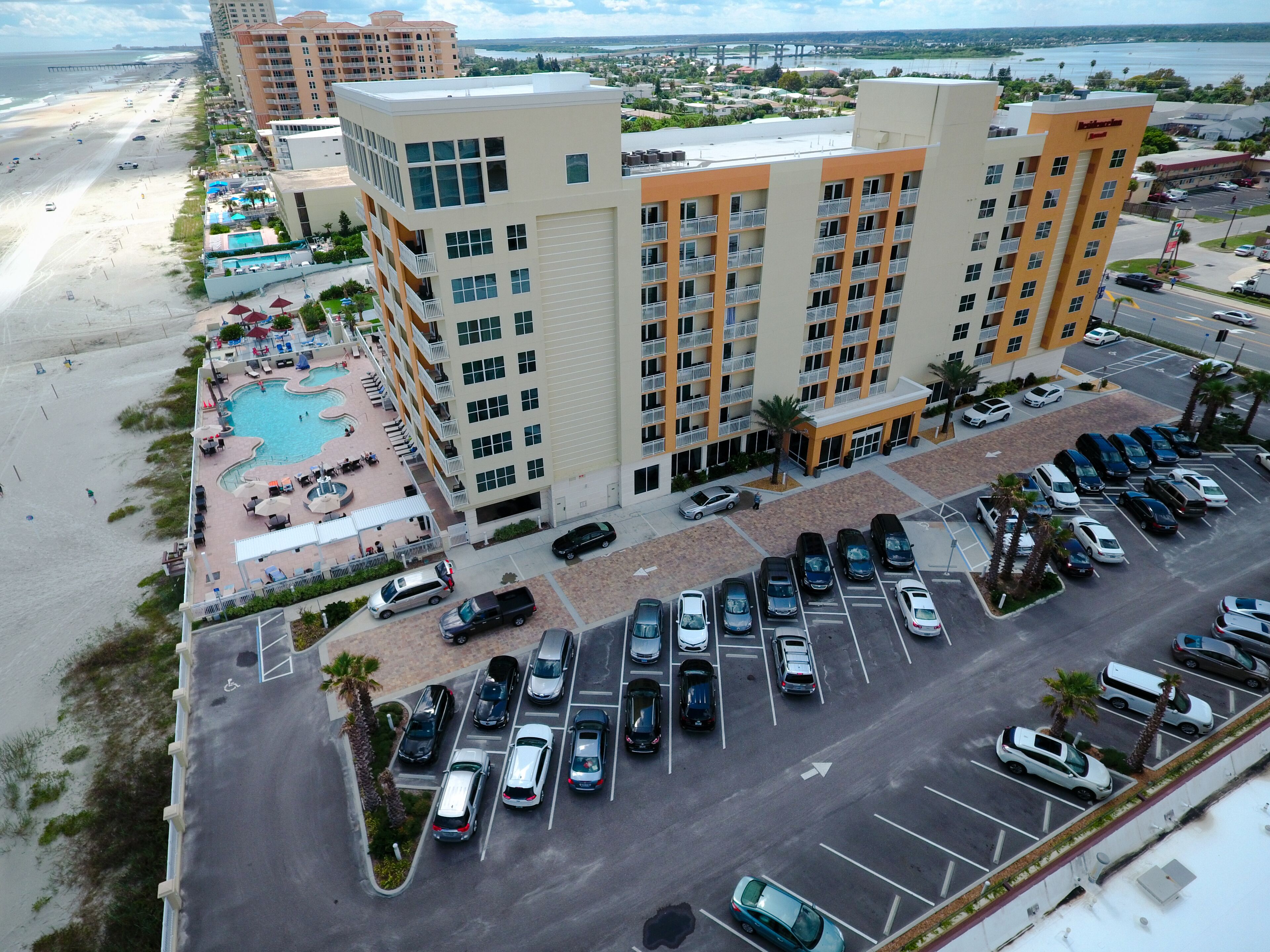 A hotel with a swimming pool, parking lot, and beachfront view.