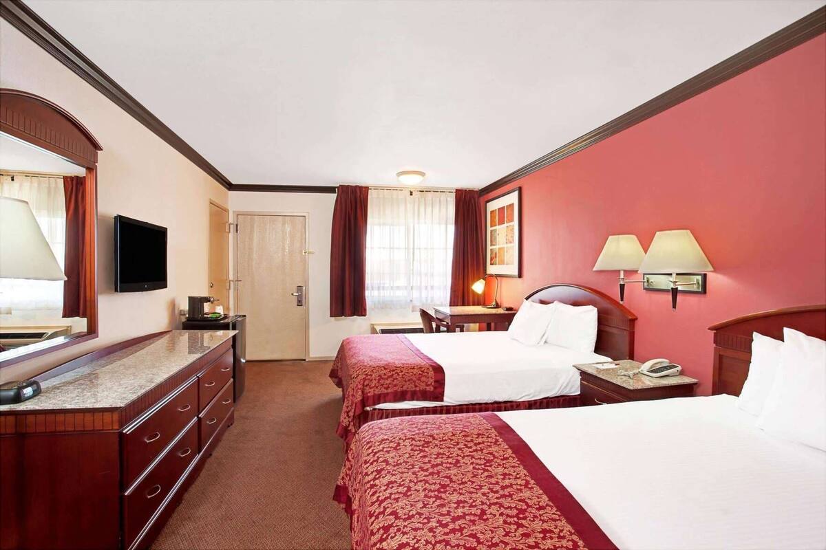 A hotel room with two beds, a desk, a chair, a television, a mirror, a dresser, and a window with curtains.