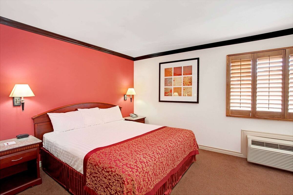 A hotel room with a bed, two bedside lamps, a framed artwork, a window with blinds, and a wall-mounted heater.