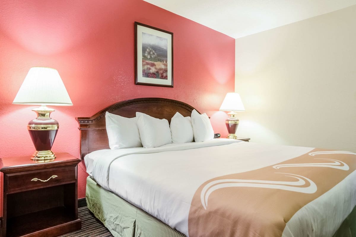 A hotel room with a large bed, two bedside tables with lamps, a framed picture on the wall, and a red accent wall.